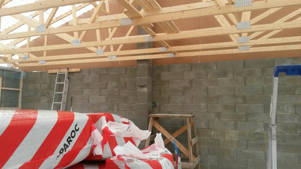 Rakennustyömaa, jossa näkyy puurakenteita, harkkoseinä, kaksi tikapuut ja PAROC-eristepaketti, valaistus on kirkas.