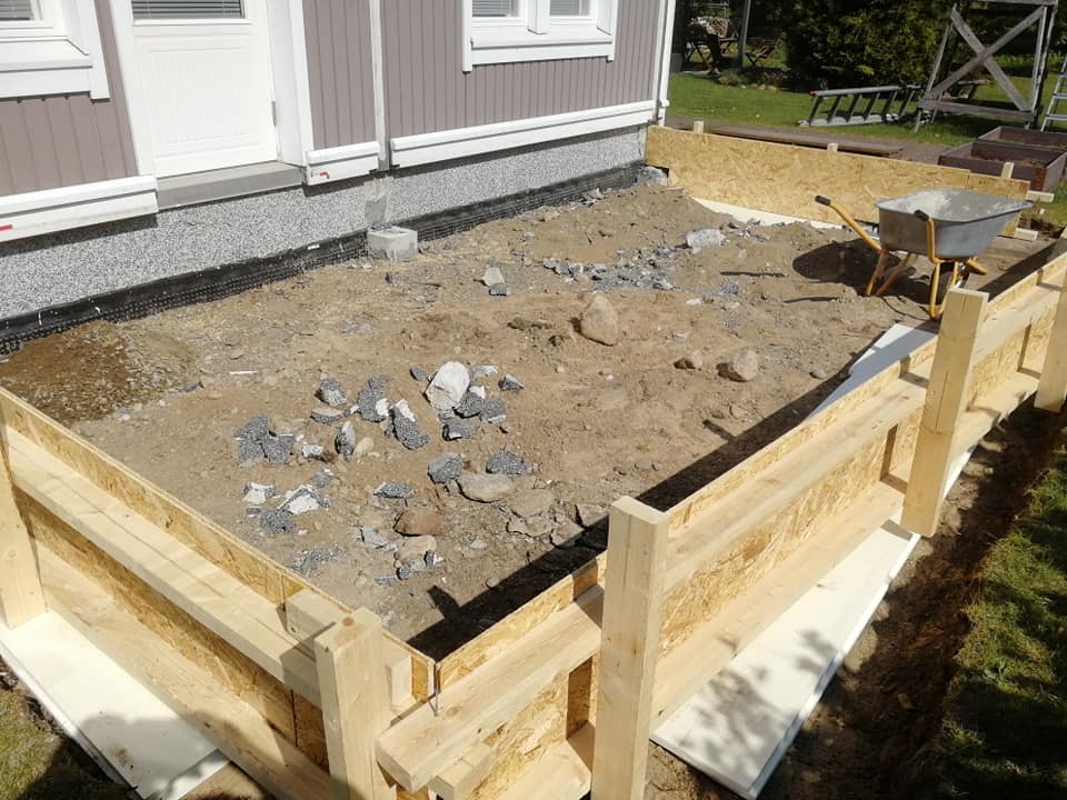 Ulkona on rakennustyömaa, jossa on puinen kehikko, maaperä, kottikärryt ja rakennusmateriaaleja, valaistus on luonnollinen.