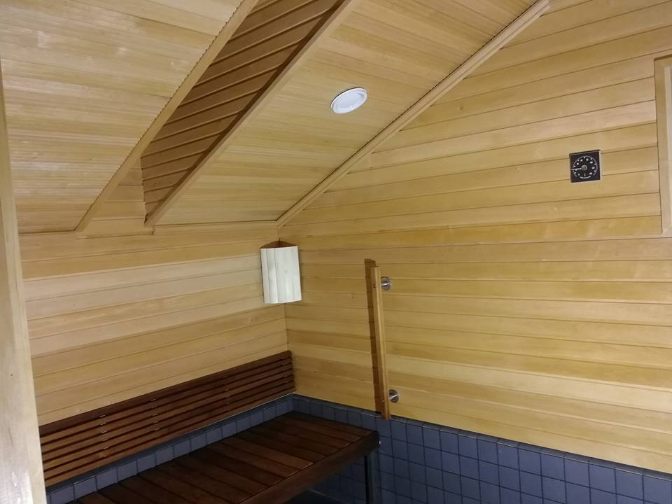 Puinen sauna, jossa on penkki, valaisin ja ilmanvaihtoaukko. Seinät ja katto ovat vaaleaa puuta, lattia on tummaa laattaa.