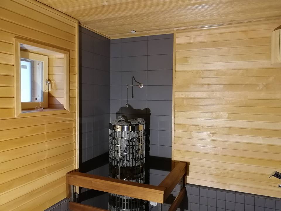 Sauna, jossa on puupanelointi, harmaat laatat, ikkuna ja kiuas kivillä.