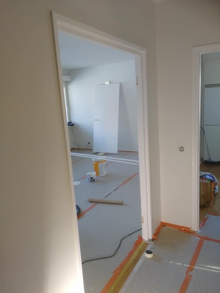 Sisätiloissa näkyy valkoiset seinät, kaksi oviaukkoa valkoisilla karmeilla, lattialla suojapaperi ja maalaustarvikkeita. Huone on valoisa.