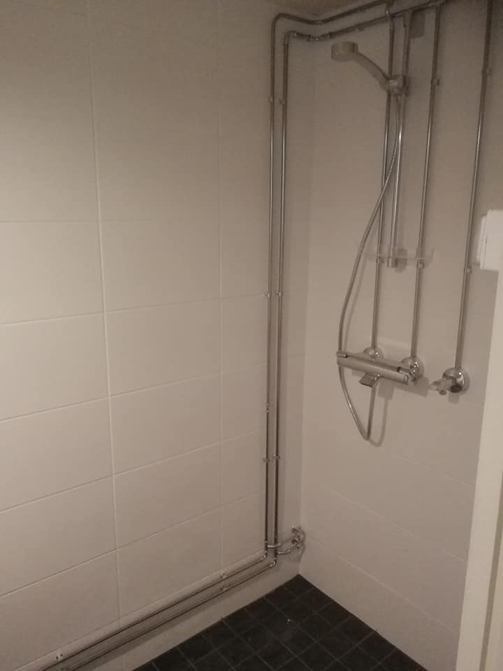 Kylpyhuoneen suihkunäkymä, jossa on valkoiset laatat, tumma lattia ja näkyvät putket.