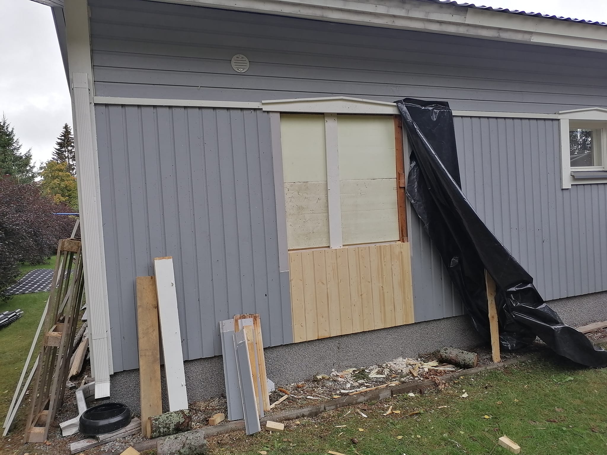 Harmaa puutalo, jossa on osittain korjattu seinä, musta suojapeite ja rakennustarvikkeita nurmikolla.