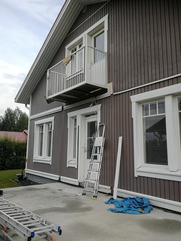 Ruskean talon ulkokuva, jossa on valkoiset ikkunat ja parveke, sekä tikkaat ja sininen pressu maassa.