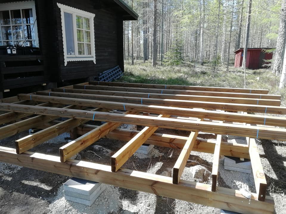 Ulkona on rakenteilla puinen terassi, jonka alla on betonilaattoja ja taustalla näkyy metsä ja punainen rakennus.