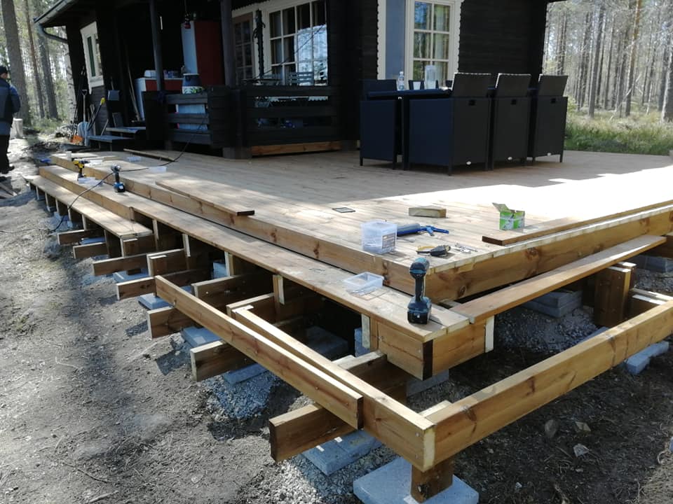 Puinen terassi rakenteilla, jossa näkyy puutavaraa, työkaluja ja osittain valmis kansi. Valoisa ulkotila.