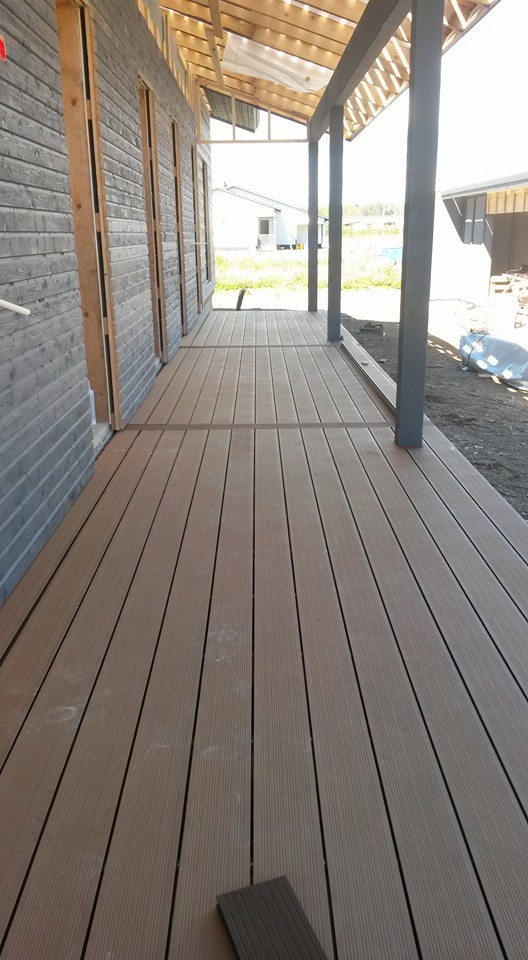 Ulkoterassi, jossa on ruskeat komposiittilaudat, harmaa seinä ja pystyt tukipylväät. Valoisa sää.