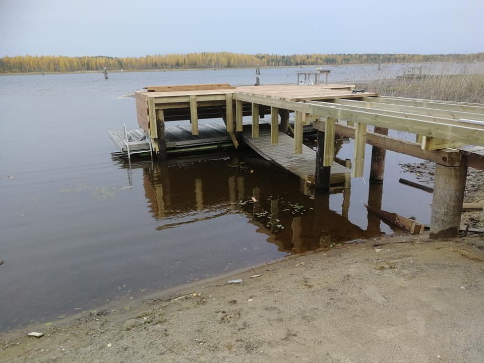 Kuva esittää uutta puista laituria, joka on osittain veden päällä ja osittain rannalla. Laituri on vaaleaa puuta ja sen rakenteet ovat selkeästi näkyvissä. Taustalla näkyy järvi ja metsää. Valo on himmeää ja pilvistä.