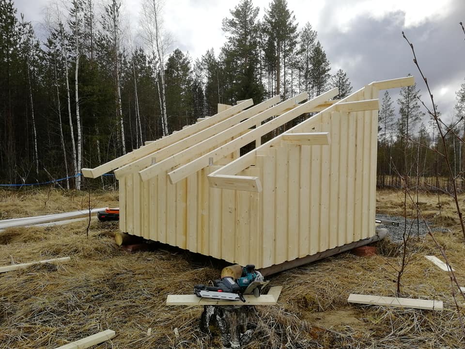 Puurakenteinen vaja rakenteilla, jossa on puinen runko ja kattopalkit, sijaitsee metsäisessä ympäristössä harmaalla säällä.