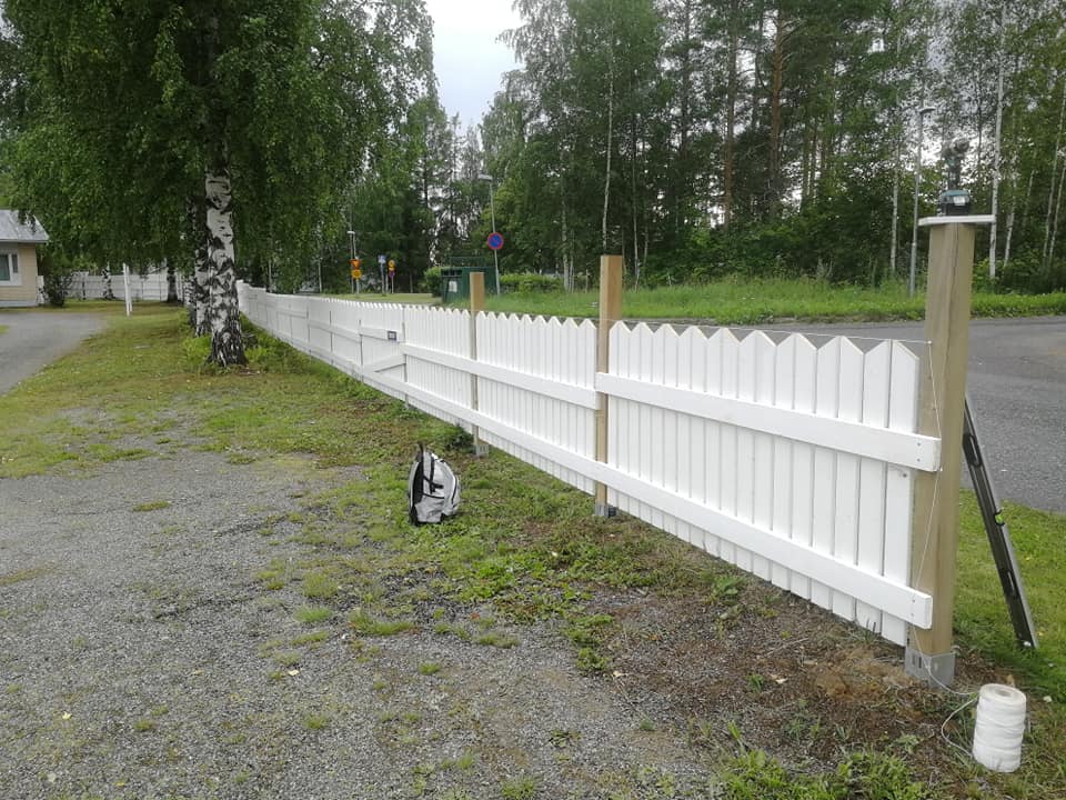 Valkoinen aita reunustaa tietä, puupylväät tukevat sitä, ja taustalla näkyy metsä ja harmaa reppu.