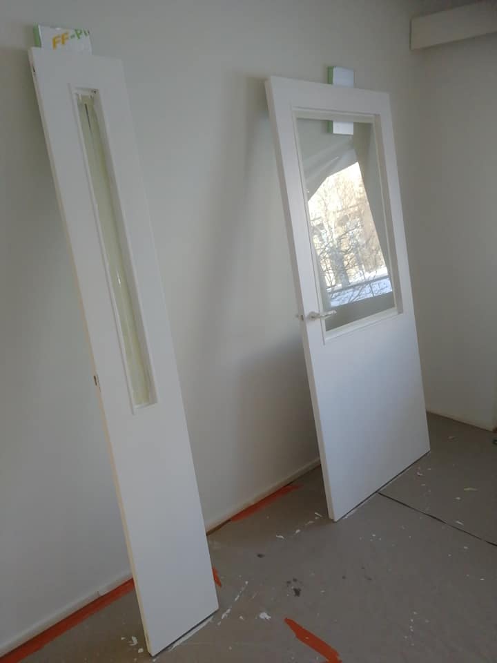 Kaksi valkoista sisäovea nojaavat valkoiseen seinään, toisessa on pitkä lasi-ikkuna ja toisessa suurempi ikkuna ja ovenkahva.